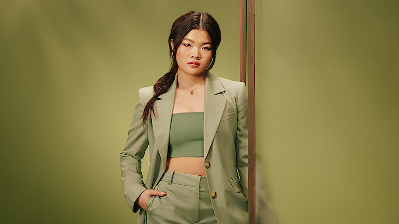 Miyako, a woman in a light green suit and crop top stands confidently against a matching green background, with one hand in her pocket, her hair styled in a loose side braid, conveying a modern, stylish atmosphere.