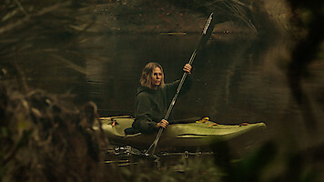 A person paddling a green kayak on a calm, forested river surrounded by trees and branches, wearing a dark hoodie and looking ahead with a serious expression, in a natural outdoor setting.