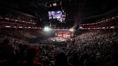 Large, packed arena hosting a WWE wrestling event with bright lights, digital screens, and enthusiastic crowd surrounding a central ring under the arena’s high ceiling.