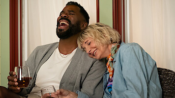Two people sitting closely together indoors, laughing and smiling. One person is holding a drink while the other leans on their shoulder, creating a relaxed and joyful atmosphere. Warm lighting and comfortable setting.