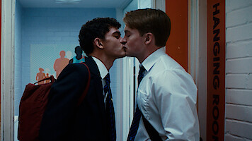 Two young men in school uniforms kiss in a hallway next to a door labeled "HANGING ROOM." One wears a backpack, and the background features colorful wall art and a white brick wall.