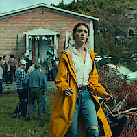 A woman in a yellow raincoat stands outside a rustic wooden house, looking concerned. Behind her, a group of people gather near the house, with a forested hillside in the background, suggesting a rural community setting.