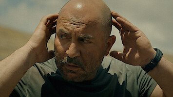 Bald man outdoors under cloudy sky, wearing a gray t-shirt and a watch, holding both hands to his head, looking concerned or focused in a natural setting.