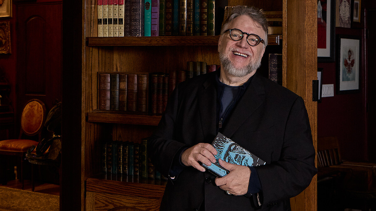 Guillermo del Toro holds a copy of the book of Frankenstein while standing in front of a book shelf.