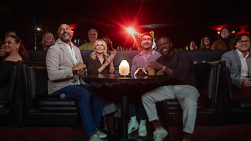 Keegan-Michael Key, Chelsea Handler, Kumail Nanjiani, and Kevin Hart seated at a round table in a dimly lit comedy club or lounge, smiling and watching a show, with a glowing candle on the table and an audience in the background.