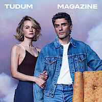 Two people stand closely together, one in a dark tank top and the other in a denim jacket, against a pastel sky backdrop with the words "TUDUM MAGAZINE" above them. An orange patterned chair is partially visible in the foreground.