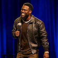 Stand-up comedian performing on stage, smiling and holding a microphone, with a dark blue curtain backdrop and stage stool partly visible.