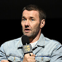 Man in a light denim jacket speaking into a microphone on stage against a dark background, appearing engaged and thoughtful in a formal event setting.
