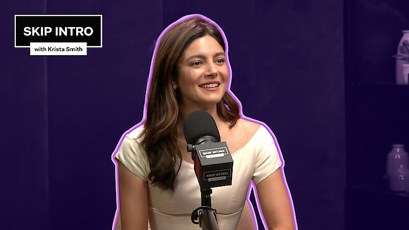 Monica Barbaro smiles in front of a microphone, framed by a deep purple background with a matching purple outline surrounding her.