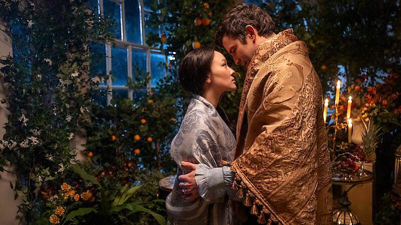 A man and woman in ornate robes share an intimate moment in a lush, candlelit indoor garden, surrounded by plants, flowers, and fruits near a large window at night.