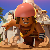 LEGO minifigure with red helmet and determined expression running on dusty ground in front of a stone structure with other minifigures and blue sky in the background.