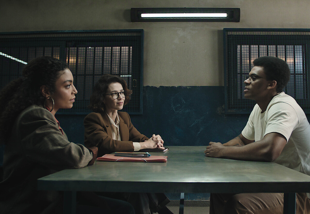 Duas mulheres, as atrizes Marjorie Estiano e Any Gabrielly, se sentam com um homem, o ator Hypólito, dentro de uma penitenciária