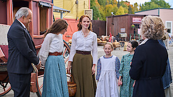 Group of people in period clothing stand and converse in a rustic, old-fashioned town setting with a stagecoach, wooden buildings, and outdoor market visible in the background.