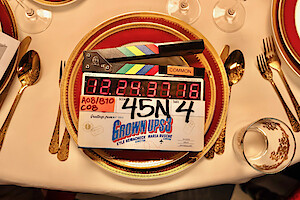 A movie clapperboard labeled "Grown Ups 3" on an elegant dining table set with gold utensils, red and gold plates, glassware, and a white tablecloth.