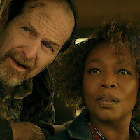 A concerned older man and a woman sit closely together inside a car, both looking intently at something outside, suggesting suspense or curiosity in a dramatic setting.