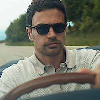 Man wearing sunglasses and a light colored shirt driving a car on a tree-lined road during the day, focused expression, countryside scenery visible in the background.
