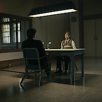 Two people sit across from each other at a table in a dimly lit interrogation room, with a large mirror on the wall reflecting the scene and a microphone on the table, creating a serious and tense atmosphere.