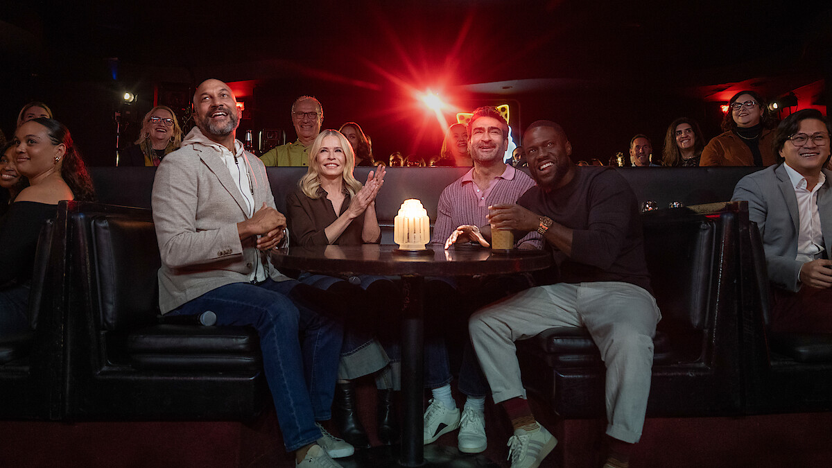Keegan-Michael Key, Chelsea Handler, Kumail Nanjiani, and Kevin Hart seated at a round table in a dimly lit comedy club or lounge, smiling and watching a show, with a glowing candle on the table and an audience in the background.