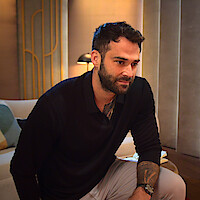 Alex Henderson in a black shirt sits in a modern, warmly lit living room with a sofa, abstract pillows, and a lamp in the background, appearing thoughtful and relaxed.