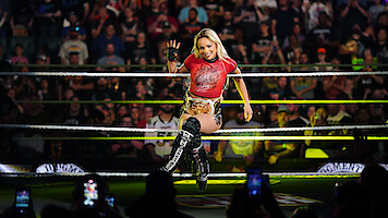 A female wrestler wearing a championship belt poses confidently in a wrestling ring, with a cheering crowd and bright arena lights creating an energetic and celebratory atmosphere. Fans are capturing the moment on their phones.