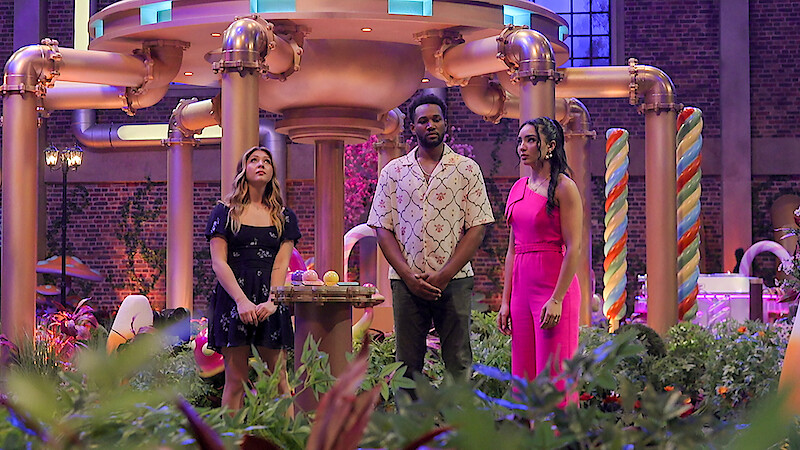 Three people stand in a whimsical, candy-themed indoor set with bright decor, oversized pipes, and colorful candy cane props, surrounded by plants and soft lighting, giving a playful, fantasy atmosphere.
