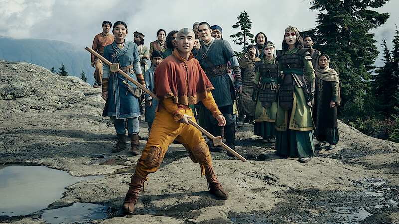 A group of people in costumes stand outdoors on rocky terrain with trees and mountains in the background, the central figure holding a staff and looking alert.