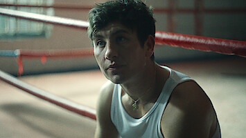 A young man in a white tank top sits inside a boxing ring, looking up thoughtfully. The gym setting has red ropes and soft, natural light coming in from a window in the background.