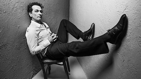 Man in formal attire sits on chair in a corner, legs extended and propped up against the walls. Black and white photo with dramatic lighting, creating a stylish, contemplative, slightly edgy mood in a minimal setting.