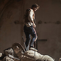 A man in rugged clothing stands atop a broken statue inside a dimly lit, dusty interior. The statue has arrows lodged in it, and the environment suggests abandonment or conflict.