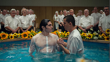 Two men in white shirts stand in a baptismal pool surrounded by sunflowers, with a group of people in white shirts and blouses watching in the background, in a brightly lit indoor setting.