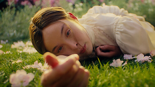 A young woman in a light blouse lies on green grass surrounded by pink flowers, looking directly at the camera in a sunlit garden or field.