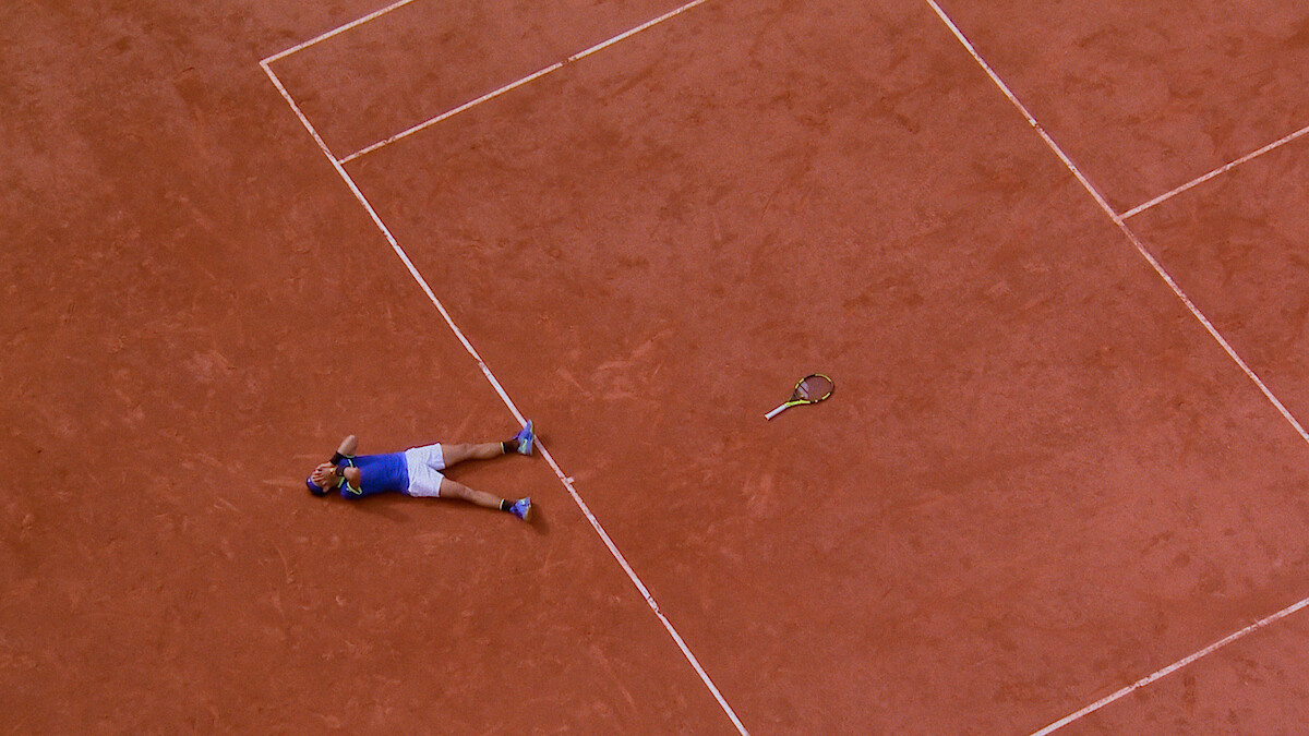 A tennis player lies on a clay court next to a tennis racquet, possibly celebrating or exhausted, viewed from above. The scene captures the lines and earthy texture of the tennis court.