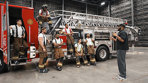 A group of firefighters in gear gather around a fire truck inside a large indoor facility while listening to a man giving instructions or a briefing.