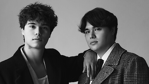 Two young men pose closely together in a studio setting, one resting his arm on the other's shoulder. Both wear jackets; the environment is minimal, and the photo is in black and white, giving a classic portrait feel.