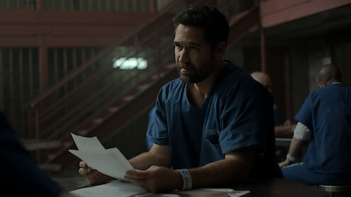 Man in blue prison uniform sits at a table holding papers in a dimly lit jail common area, with other inmates in the background and metal staircases visible.