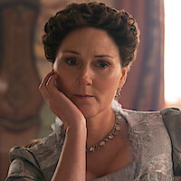 A woman with styled brown hair and vintage clothing sits indoors on a chair, resting her head on her hand, with soft natural light and ornate décor in the background, evoking a historical or period setting.