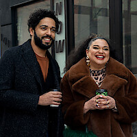 Two people standing outside a building, smiling and holding coffee cups; the man wears a dark coat, and the woman is dressed in a brown fur coat with statement earrings. Urban setting, casual atmosphere.