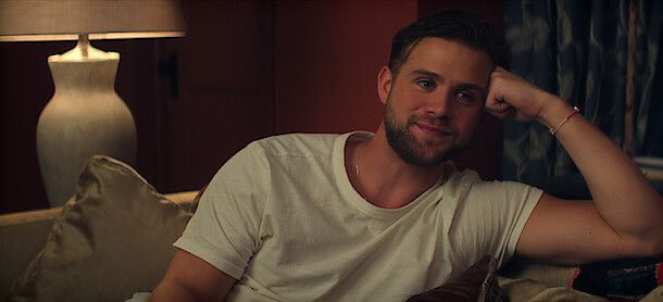Leo Woodall as Vladimir in a white t-shirt relaxing on a sofa in a cozy, dimly lit living room with pillows, a lamp, and curtains in the background.
