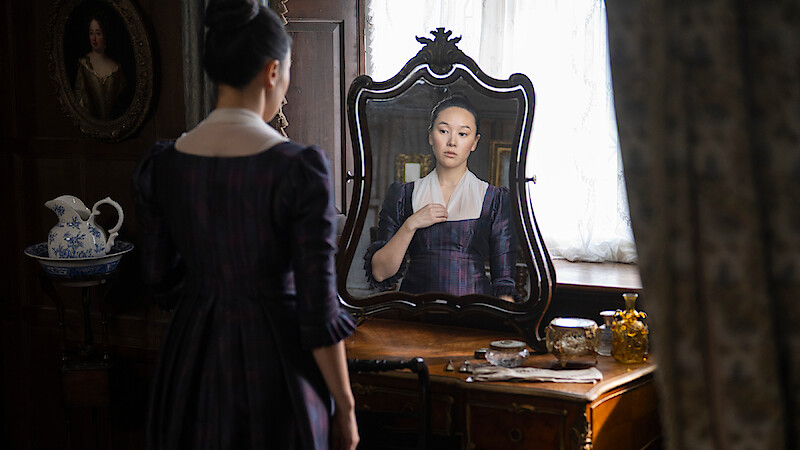 A woman in historical clothing stands at a wooden vanity, looking at her reflection in an ornate mirror, with antique items and soft natural light filling the elegant, vintage room.