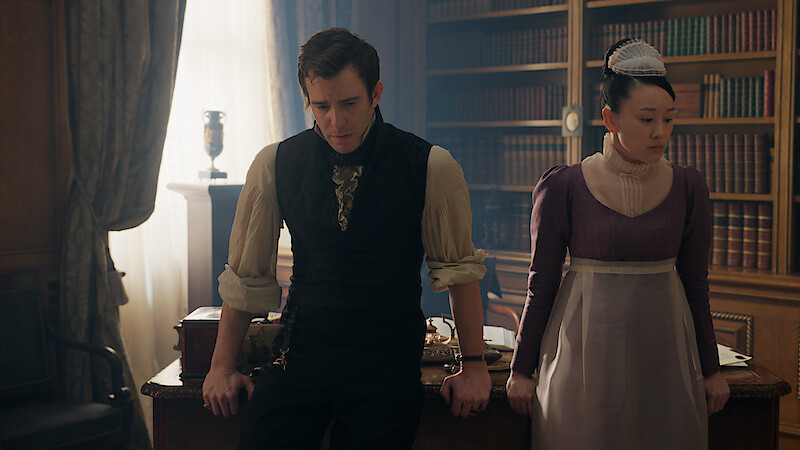 A man and woman in historical attire lean against a desk in a richly furnished study with bookshelves, draped curtains, and warm lighting, suggesting a period drama setting.