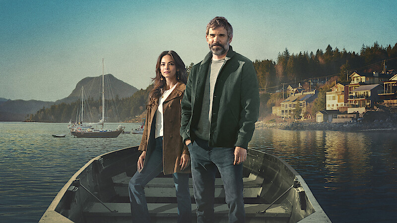 Man and woman standing in a rowboat on calm water, with sailboats, mountains, and a small waterfront town in the background. The setting appears peaceful, natural, and scenic.