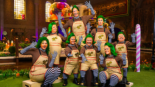 Group of people dressed as Oompa Loompas with green wigs, brown shirts, striped leggings, and overalls in a colorful, whimsical candy factory setting with oversized sweets and bright lighting.