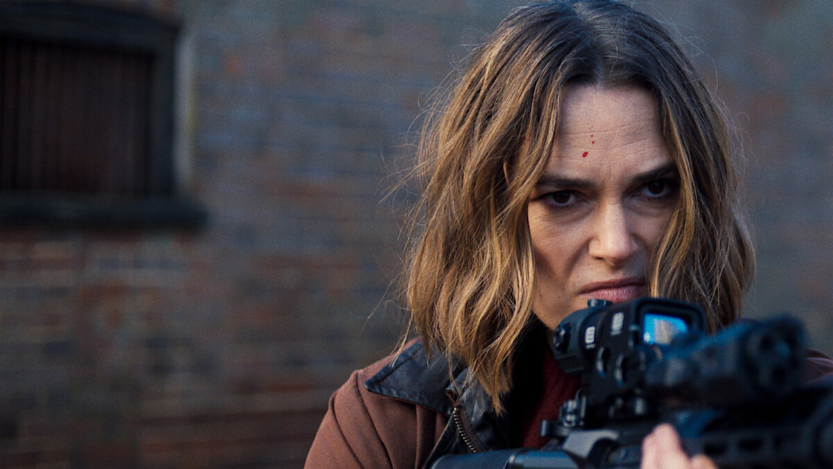 Woman with shoulder-length hair aiming a rifle, looking intensely forward, standing outdoors near a brick wall, focused and alert in an urban environment.