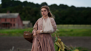 Woman in a long, old-fashioned dress with an arm in a sling walks outdoors near a field, holding a wicker basket. A rustic farmhouse and green trees are visible in the background, suggesting a rural historical setting.