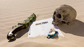 A sword, a human skull, a compass, and a ‘ONE PIECE’ script page arranged on desert sand, evoking an adventurous, mysterious, and slightly ominous atmosphere.