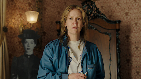 A concerned person in a blue jacket stands in a vintage room with patterned wallpaper, ornate wooden furniture, and a black-and-white cutout of a woman in period clothing in the background.