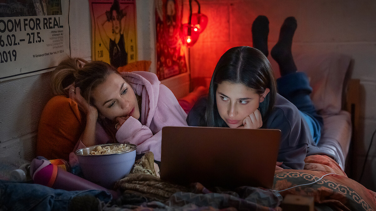 Two young women lying on a bed in a cozy, dimly lit room, watching a laptop together. There is a bowl of popcorn, posters on the wall, and warm blankets, creating a relaxed, intimate atmosphere.
