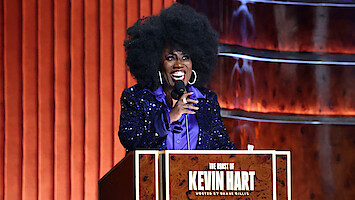  Sheryl Underwood with a large afro hairstyle and sparkly purple jacket speaks at a podium labeled "The Roast of Kevin Hart," on a stage with wood paneling and warm lighting.