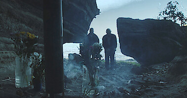 In the foreground, cut flowers and burning incense; in the background two figures visible beyond a rock formation.
