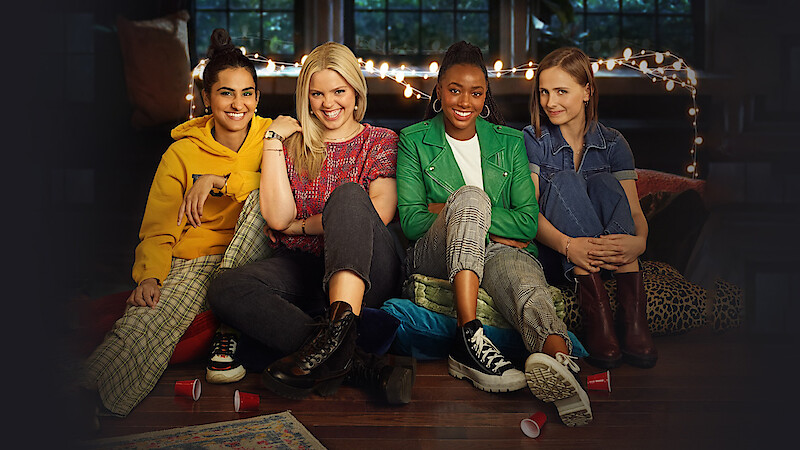 Four young women sit on the floor together in a cozy room with string lights and large windows, smiling and relaxed, surrounded by pillows and cups, suggesting a casual indoor gathering or hangout.